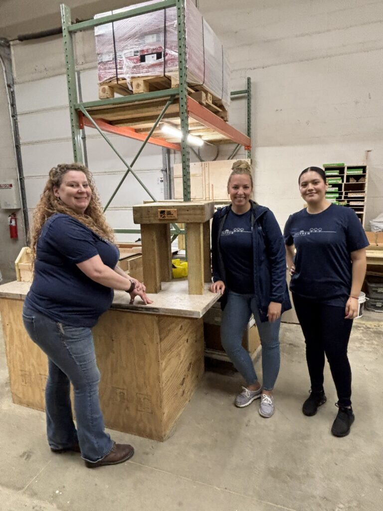 Three Intandem Credit Union volunteers standing beside handcrafted logoed tables built during a community involvement project with Home Repair Services.